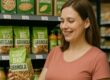 Frau mit Kind im Supermarkt vor Regal mit Bio- und Vegan-Granola, freut sich über gesunde Produkte – BRIZZLY Villach Coaching